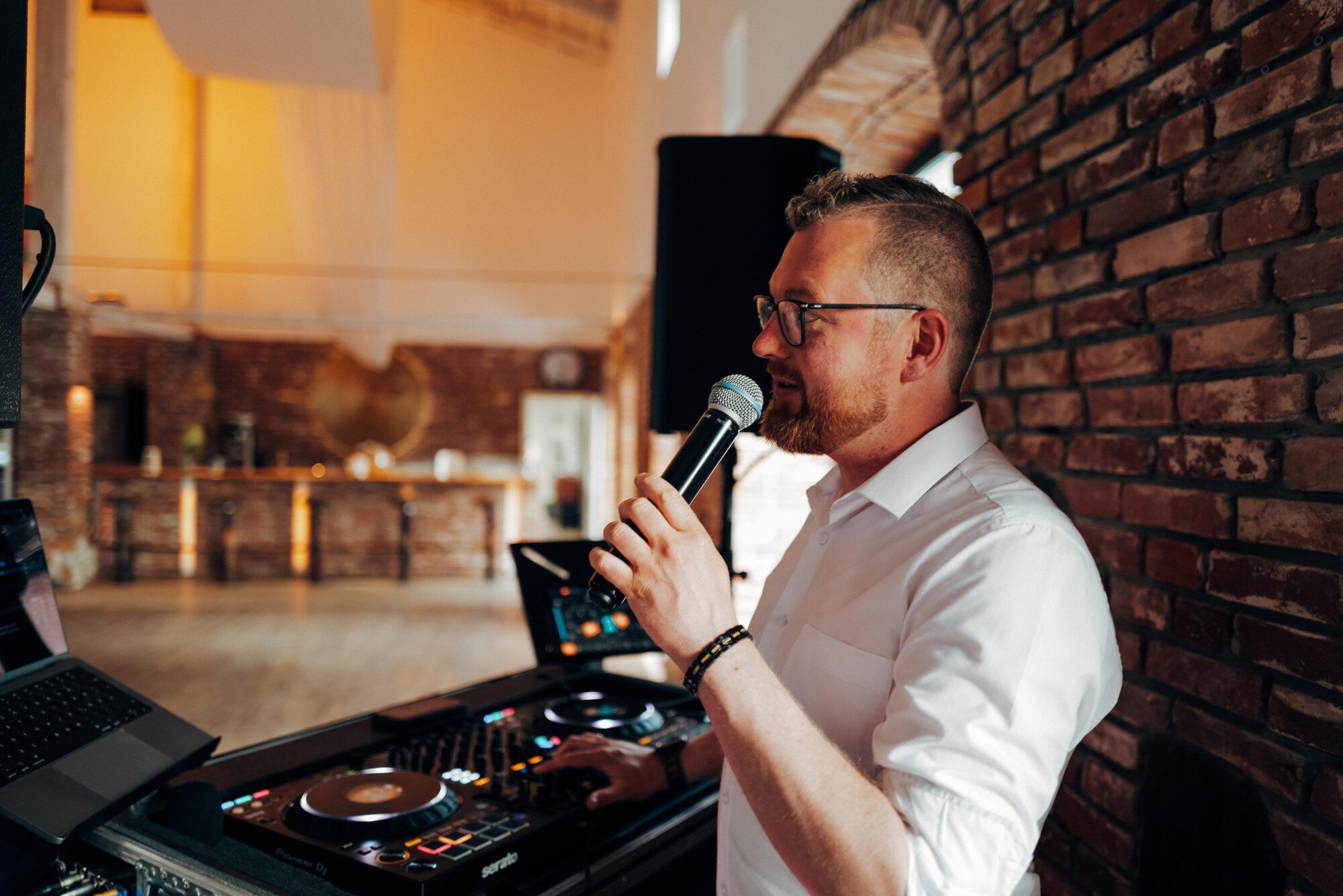 Dennis Genske moderiert mit Mikrofon am DJ-Pult in einer Hochzeitsscheune